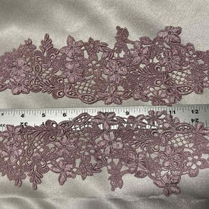 May include: Two mauve-colored floral lace appliques. Each applique features a detailed design of flowers and leaves. A metal ruler is placed across the appliques, showing measurements in inches. The appliques are likely for sewing or crafting projects.