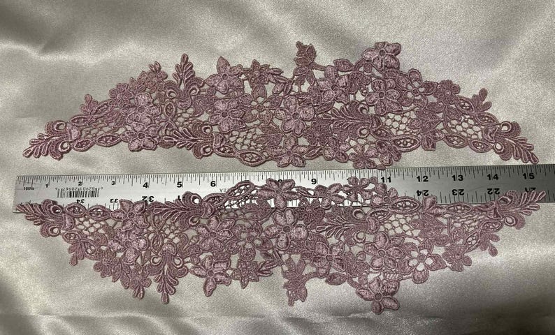 May include: Two mauve-colored floral lace appliques. Each applique features a detailed design of flowers and leaves. A metal ruler is placed across the appliques, showing measurements in inches. The appliques are likely for sewing or crafting projects.
