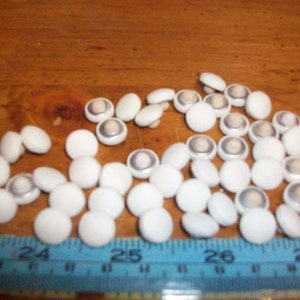 Puede incluir: Un montón grande de botones blancos con un centro plateado. Los botones están dispuestos en un patrón aleatorio sobre una superficie de madera.