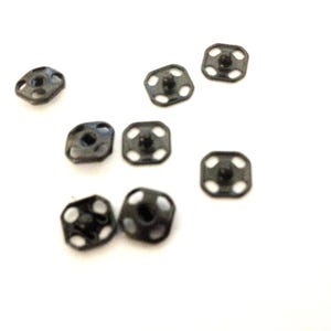 Puede incluir: Primer plano de diez botones a presión de metal negro. Los botones a presión son de forma cuadrada con cuatro agujeros alrededor del borde y un centro elevado.