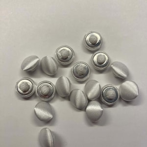 May include: A collection of small, white, fabric-covered buttons with a silver metal rim and a central, raised, circular design. The buttons are scattered across a white surface, showcasing their delicate texture and design.