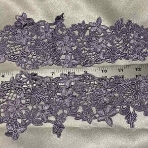 May include: Two lavender floral lace appliques with intricate flower and leaf designs. A ruler shows the appliques are approximately 16 inches long. The lace is detailed and suitable for various craft projects.