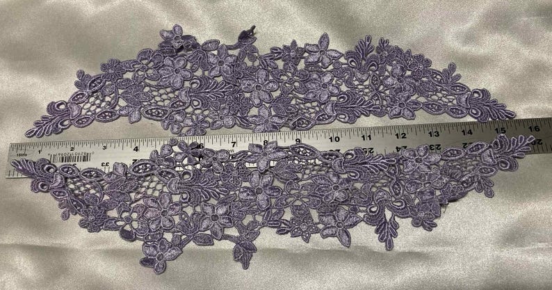 May include: Two lavender floral lace appliques with intricate flower and leaf designs. A ruler shows the appliques are approximately 16 inches long. The lace is detailed and suitable for various craft projects.