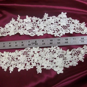 May include: Two pieces of ivory floral lace trim with a ruler for scale. The lace features a delicate, openwork design with various flower and leaf shapes. The trim is displayed on a deep burgundy fabric.