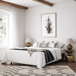 May include: A bedroom scene featuring a large white bed with decorative pillows and a dark gray throw blanket. A framed artwork with a gold frame hangs above the bed. The room has a neutral color palette with a patterned rug.