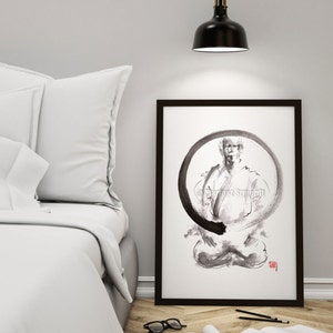 May include: Black framed art print featuring a seated figure within a black circular design. The artwork is in black and white, with the artist's signature in the upper right corner. The print is displayed on a wooden floor next to a bed.
