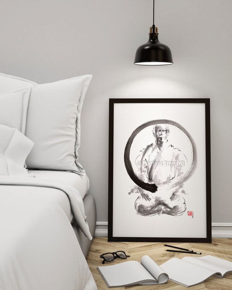May include: Black framed art print featuring a seated figure within a black circular design. The artwork is in black and white, with the artist's signature in the upper right corner. The print is displayed on a wooden floor next to a bed.