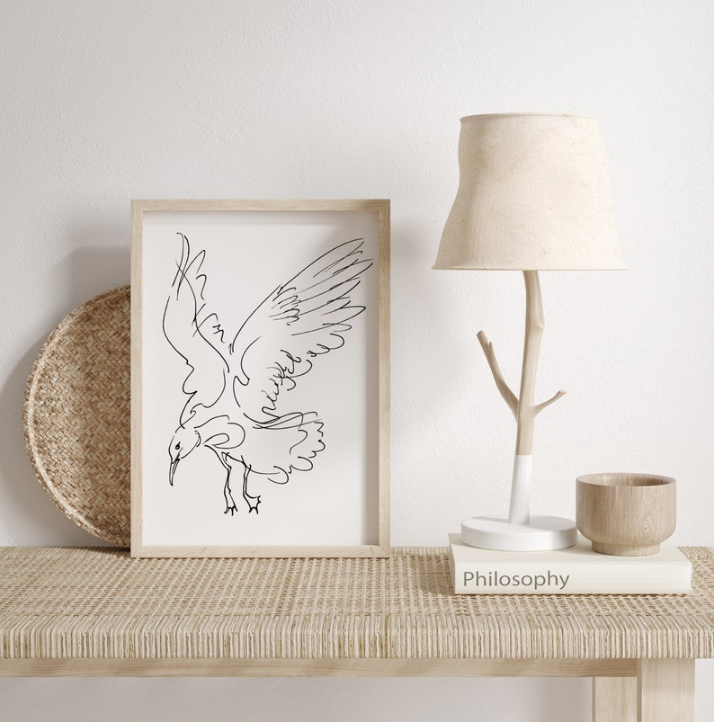 May include: A framed art print featuring a minimalist black line drawing of a bird in flight. The artwork is displayed on a woven table, alongside a beige lampshade, a wooden bowl, and a book titled "Philosophy".