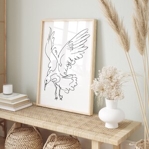 May include: A framed art print featuring a minimalist line drawing of a bird in flight. The artwork is in black against a white background, encased in a light wood frame. The print is displayed on a woven bench, alongside decorative elements.