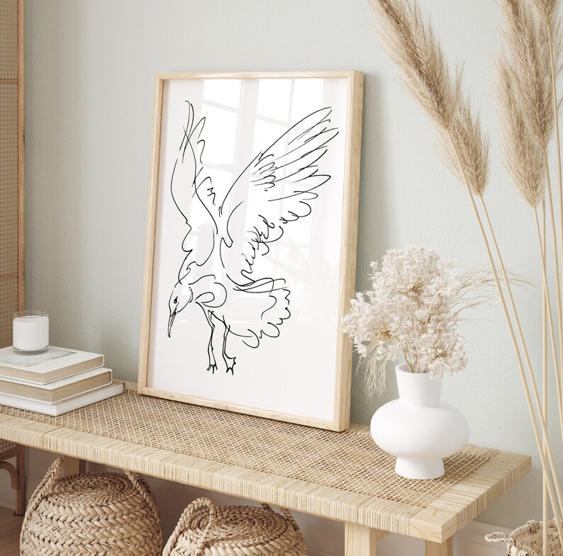 May include: A framed art print featuring a minimalist line drawing of a bird in flight. The artwork is in black against a white background, encased in a light wood frame. The print is displayed on a woven bench, alongside decorative elements.