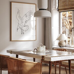 May include: A dining room scene featuring a framed black and white line drawing of a bird in flight. The room includes a rectangular table with a marble-like top, rattan chairs, and a white pendant light. A window with a bamboo shade and a table lamp are also visible.