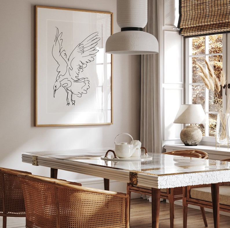 May include: A dining room scene featuring a framed black and white line drawing of a bird in flight. The room includes a rectangular table with a marble-like top, rattan chairs, and a white pendant light. A window with a bamboo shade and a table lamp are also visible.