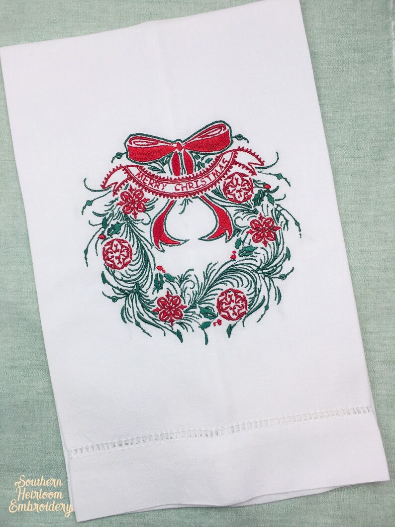 以下が含まれることがあります： 赤いリボンと「メリークリスマス」の文字が刺繍された、赤と緑のクリスマスリースが描かれた白いキッチンタオルです。