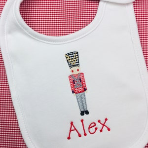 May include: White baby bib with a red and blue embroidered toy soldier and the name Alex in red.