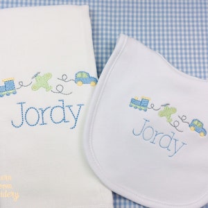 May include: A white burp cloth and bib with blue and yellow embroidered vehicles, a plane, and the name "Jordy".