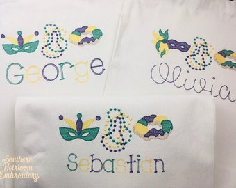 May include: Three white towels with embroidered Mardi Gras themed designs. The towels feature a purple, green, and gold mask, beads, and a king cake. The names George, Olivia, and Sebastian are embroidered on each towel.