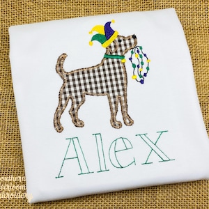 May include: White embroidered t-shirt with a brown and white checkered dog wearing a green, yellow, and purple jester hat and holding a string of purple, green, and yellow beads. The name "Alex" is embroidered in green below the dog.