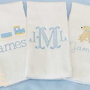 May include: Three white baby burp cloths with embroidered designs. One cloth features a blue train and the name "James". Another has a blue monogram "M". The third cloth displays a tan dog with a blue bow and the name "James".