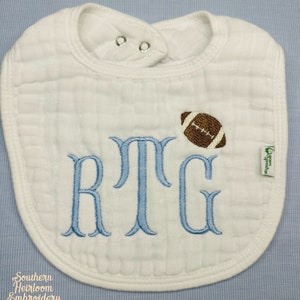 May include: White baby bib with blue embroidered monogram "RTG" and a brown football graphic. The bib has two snaps at the neck.
