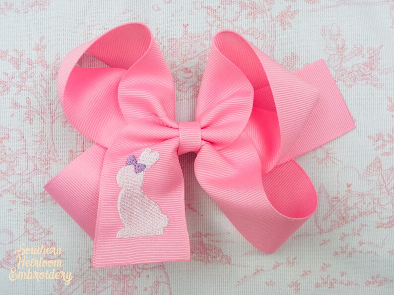 May include: A pink hair bow with a white embroidered bunny design. The bow is made of a soft, textured fabric.