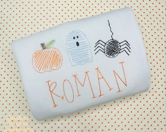 May include: White rectangular fabric with embroidered Halloween-themed designs. Features an orange pumpkin, a light blue ghost, and a black spider. The name "ROMAN" is embroidered in orange below the designs.