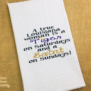 May include: White kitchen towel with embroidered text that reads "A true Louisiana woman is a Tiger on Saturdays and a Saint on Sundays!"