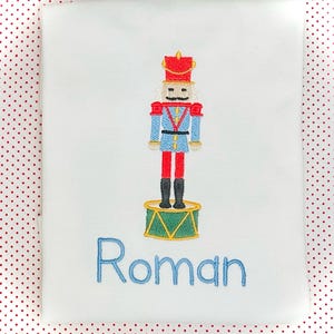 May include: A white embroidered onesie with a blue and red nutcracker design and the name "Roman" embroidered in blue below the design.