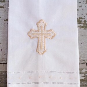 May include: White linen towel with an embroidered gold cross. The towel features a decorative border with small embroidered details. The cross is centered on the towel, and the design is simple and elegant.