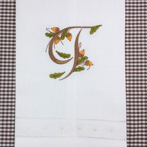 May include: White hand towel with a brown and green embroidered letter "T" with acorns and leaves. The towel has a white scalloped edge with small white dots.