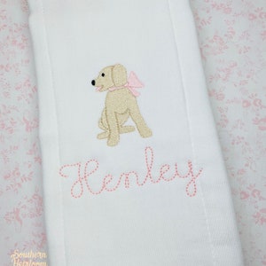 May include: White burp cloth with pink embroidery of a dog wearing a pink bow and the name Henley.