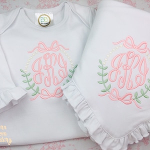 May include: A white long-sleeved onesie and a white baby blanket with a pink and green embroidered monogram in a wreath design. The monogram reads "AMY".