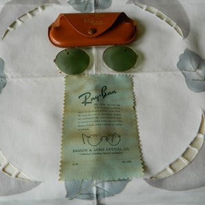 May include: Vintage Ray-Ban sunglasses with green lenses and a gold metal frame. The sunglasses are in a brown leather case and come with a Ray-Ban cleaning cloth. The cleaning cloth has the text "Ray-Ban" and "Bausch & Lomb Optical Co. "America's Leading Optical Facilities" printed on it.