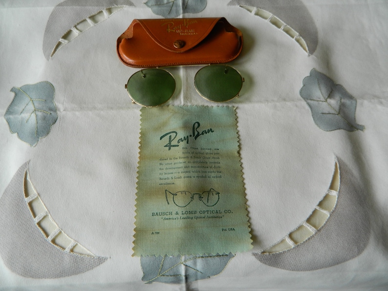 May include: Vintage Ray-Ban sunglasses with green lenses and a gold metal frame. The sunglasses are in a brown leather case and come with a Ray-Ban cleaning cloth. The cleaning cloth has the text "Ray-Ban" and "Bausch & Lomb Optical Co. "America's Leading Optical Facilities" printed on it.