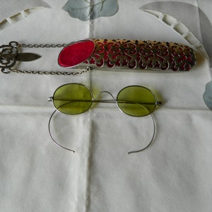 Op de afbeelding: Een paar ovale, groen getinte brillen met een zilveren montuur en een zilveren ketting bevestigd aan een decoratieve zilveren etui met een rode fluwelen voering.