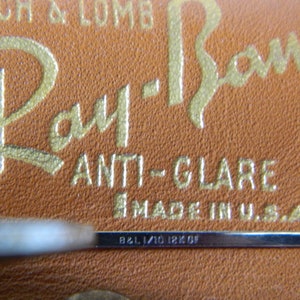 May include: Close-up of a brown leather case with gold lettering that reads "Ray-Ban ANTI-GLARE MADE IN U.S.A." and a silver metal bar with the inscription "B&L 1/10 18K OF".