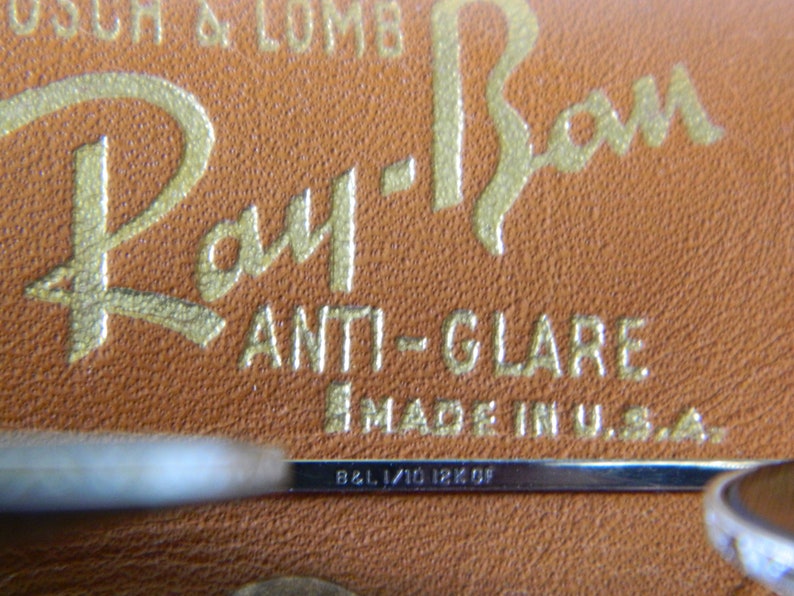 May include: Close-up of a brown leather case with gold lettering that reads "Ray-Ban ANTI-GLARE MADE IN U.S.A." and a silver metal bar with the inscription "B&L 1/10 18K OF".