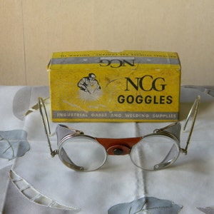 Puede incluir: Gafas de seguridad vintage NCG en una caja amarilla con el texto "NCG Goggles Industrial Gases and Welding Supplies". Las gafas son plateadas con una correa de cuero marrón y lentes transparentes.