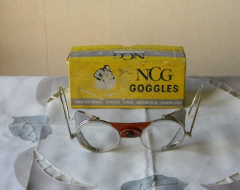 May include: Vintage NCG Goggles in a yellow box with the text "NCG Goggles Industrial Gases and Welding Supplies". The goggles are silver with a brown leather strap and clear lenses.