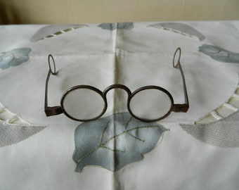 May include: A pair of antique round-framed glasses with metal frames and clear lenses. The glasses are lying on a white tablecloth with a floral pattern.