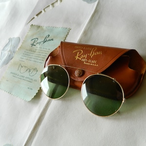 May include: Vintage Ray-Ban sunglasses with a brown leather case. The sunglasses have round green lenses and a gold metal frame. The case has the Ray-Ban logo and the text "Anti-Glare Made in U.S.A."