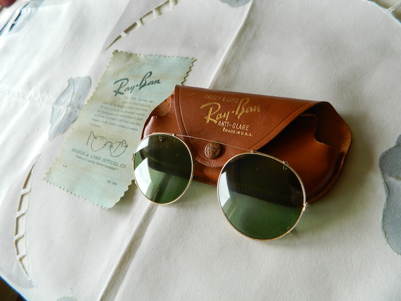 May include: Vintage Ray-Ban sunglasses with a brown leather case. The sunglasses have round green lenses and a gold metal frame. The case has the Ray-Ban logo and the text "Anti-Glare Made in U.S.A."
