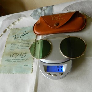 May include: Vintage Ray-Ban sunglasses with green lenses and a gold metal frame. The sunglasses are on a digital scale that reads 10.7. The sunglasses are in a brown leather case with the Ray-Ban logo.