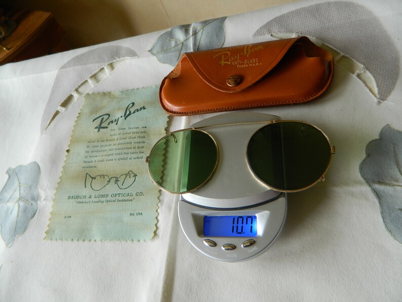 May include: Vintage Ray-Ban sunglasses with green lenses and a gold metal frame. The sunglasses are on a digital scale that reads 10.7. The sunglasses are in a brown leather case with the Ray-Ban logo.