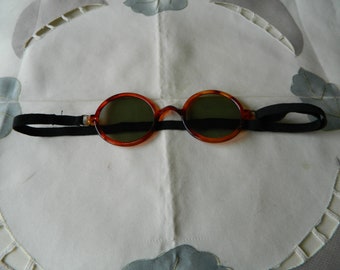 May include: Round sunglasses with a tortoiseshell frame and dark green lenses. The glasses have a black elastic strap for a secure fit. The eyewear is set against a white background with a floral pattern.