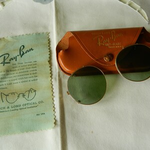 May include: Vintage Ray-Ban sunglasses with a brown leather case. The case has the text "Ray-Ban Anti-Glare" printed on it. The sunglasses have round green lenses.