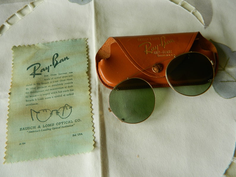 May include: Vintage Ray-Ban sunglasses with a brown leather case. The case has the text "Ray-Ban Anti-Glare" printed on it. The sunglasses have round green lenses.