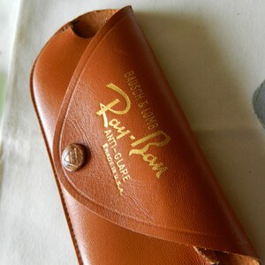 May include: A brown leather Ray-Ban sunglasses case with gold lettering that reads "Ray-Ban Anti-Glare Made in U.S.A. Bausch & Lomb".
