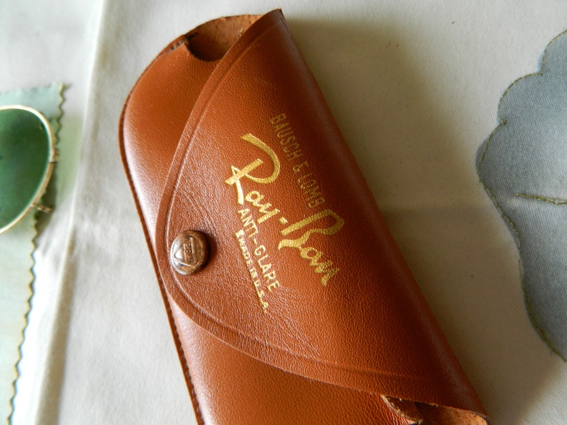 May include: A brown leather Ray-Ban sunglasses case with gold lettering that reads "Ray-Ban Anti-Glare Made in U.S.A. Bausch & Lomb".