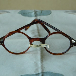 May include: Round-framed eyeglasses with a tortoiseshell pattern. The glasses have a classic design with a bridge and temples in a similar pattern. The frames are accented with silver-colored details at the hinges. A small tag with text is attached to the frame.