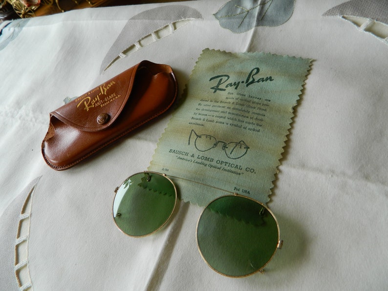 May include: Vintage Ray-Ban sunglasses with green lenses and a gold metal frame. The sunglasses are in a brown leather case with the Ray-Ban logo. The case is on a white surface with a piece of paper with the Ray-Ban logo and the text "Bausch & Lomb Optical Co. America's Leading Optical Institution."
